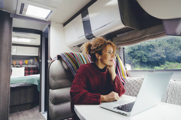 Adult woman use laptop computer inside a camper van recreational vehicle sitting at the table with bedroom in background and nature park outside the windos. Concept of travel and remote worker female people stock photo