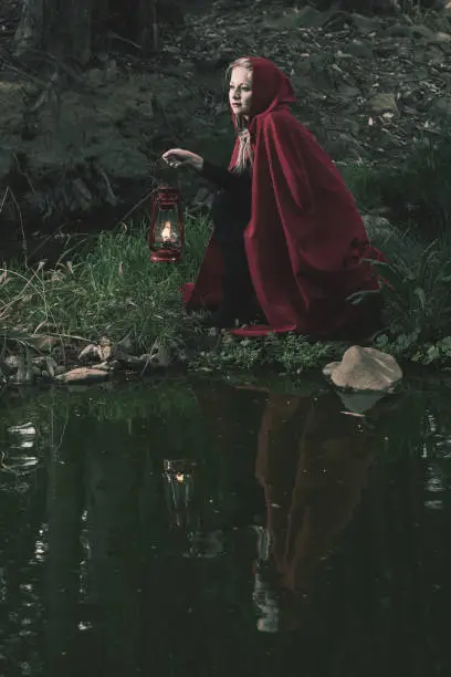 Attractive woman dressed a little red riding-hood cross a stream in a dark forest with a lantern