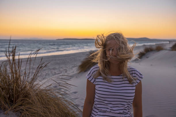 Blonde woman standing on the beach with wind in her hair at sunset, Sardinia, Italy.