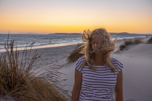 Blonde woman standing on the beach with wind in her hair at sunset, Sardinia, Italy.