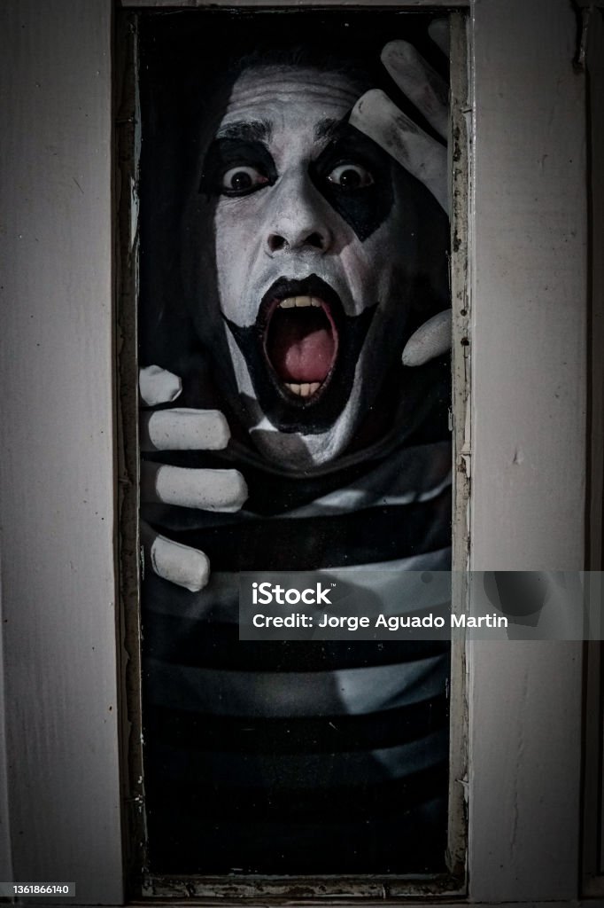 Vertical terrifying shot of a male in a mime costume posing in horror behind the window of an old rusty door A vertical shot of a male in a mime costume posing in horror behind the window Blood Stock Photo