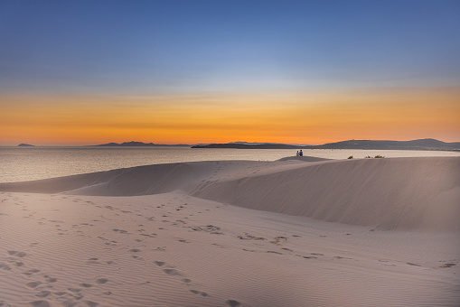 View of a couple sitting on the beach among sand dunes looking at sunset, Sardinia, Italy.