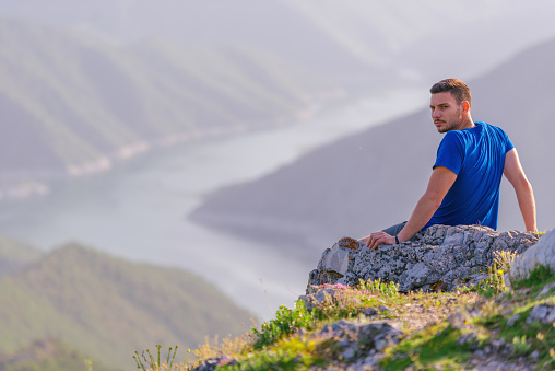 Adventurous man sitting on top of a mountain and enjoying the beautiful view, while looking downhill at the blue river and amazing mountain line.