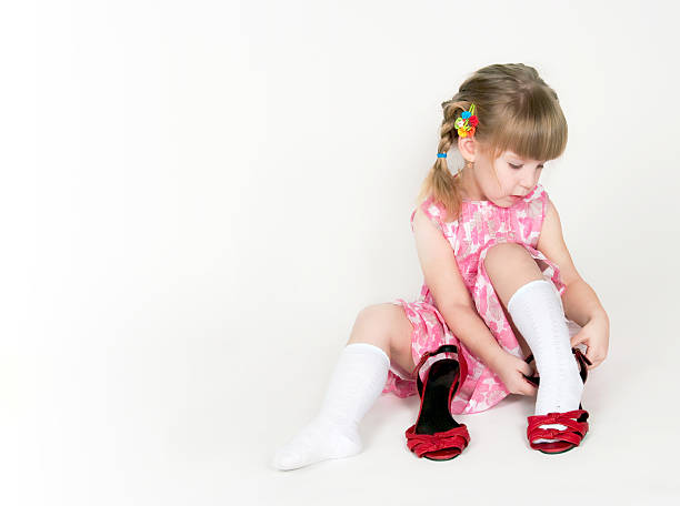 Adorable toddler girl sitting on grass, wearing a diaper and a pink dress, playing with her shoe and sticking her tongue out