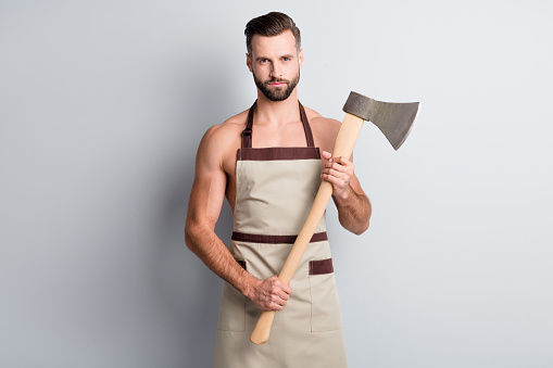 Portrait of attractive fit content athletic guy wearing apron holding in hand wooden ax isolated over light grey pastel color background.