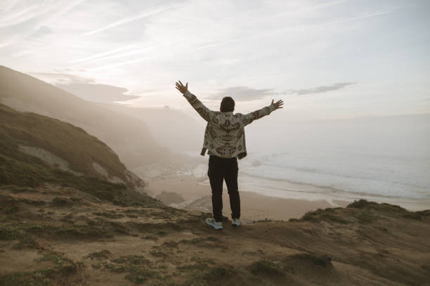 istock Young man with arms raised by the sea at sunset 1361915068