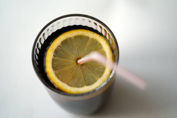 Top view of glass of cola with straw, lemon slice and ice cube on white background.