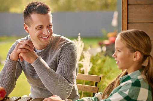 Interesting story. Happy middle-aged man in gray sweater listening attentively looking at talking smiling school-age girl sitting at table on open terrace at home on fine afternoon