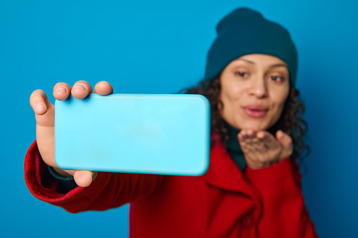 Focus on the hand of a blurred charming woman in bright red warm cozy woolen coat and green hat sending air kisses while making selfie and talking video call against blue background with copy ad space