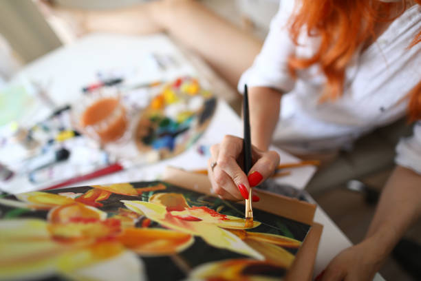 Top view of beautiful woman creating masterpiece in studio. Talented pretty artist painting picture using paintbrush. Red haired painter sitting in workshop. Art and creativity concept