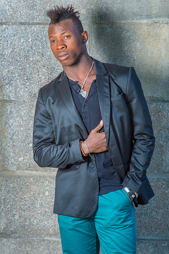 Dressing in a black blazer, green pants,  a hand in his pocket, a hand putting on his chest, a young black guy with mohawk hair is standing against the wall, looking forward.