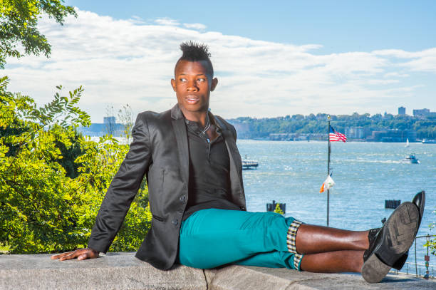 a young black guy with mohawk hair is sitting on a rocky stone by a river, stretching his legs, relaxing - mohawk river imagens e fotografias de stock