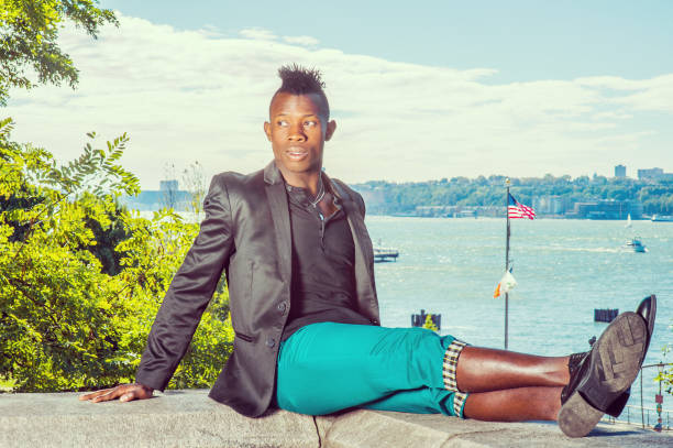 un joven negro con cabello mohawk está sentado en una cerca de piedra junto a un río, estirando las piernas, relajándose - mohawk river fotografías e imágenes de stock