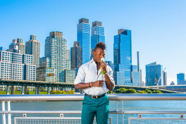 un joven negro con cabello mohawk está parado en el frente de los edificios, con las manos sosteniendo una rosa blanca, oliendo. - mohawk river fotos fotografías e imágenes de stock