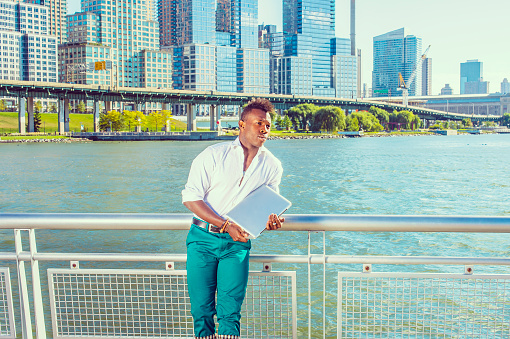 Looking for Success. Dressing in a white shirt, green pants, a young black guy with mohawk hair is standing in the front of high buildings, holding a laptop computer, thinking.