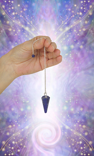 female hand holding lapis lazuli dowsing pendant against beautiful sparkling ethereal purple energy background and copy space for messages