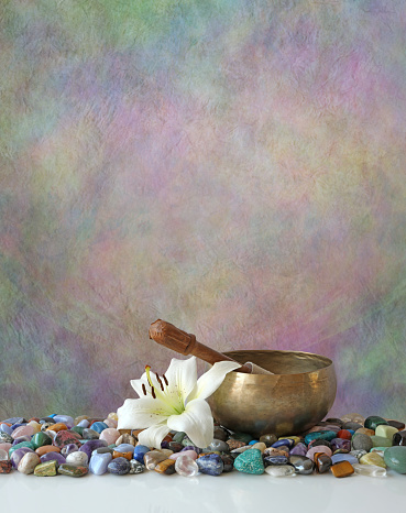 a rustic stone multicoloured background with a singing bowl sitting on tumbled healing stones and a single lily head with copy space for message