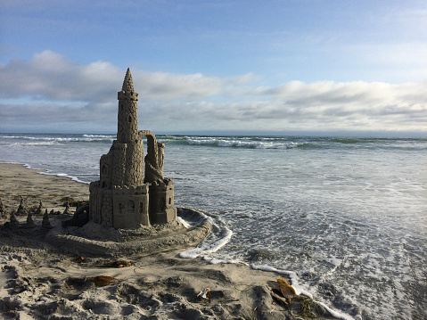 Moat protects kids' sand castle against ocean waves on a sunny day, includes blue sky and rain clouds in the background.