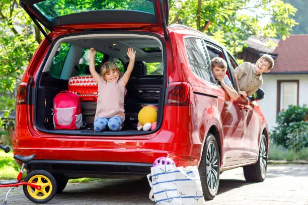 Three children, two boys and preschool girl sitting in car before leaving for summer vacations with parents. Happy kids, siblings, brothers and sister with suitcases and toys going on family road trip.