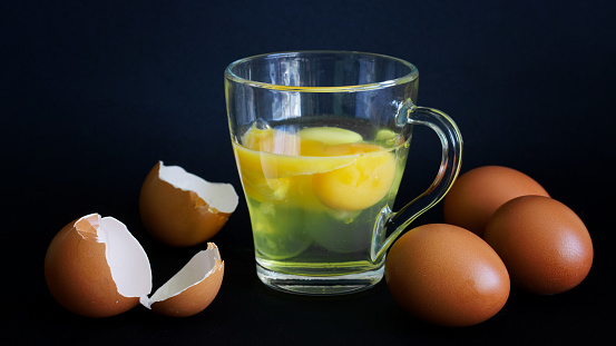 Raw chicken eggs in a transparent mug on a dark background. Nearby lies a shell of chicken eggs. Natural food without cooking. Daylight