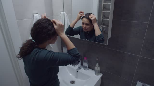Concerned woman noticing hair problem looking at mirror in bathroom
