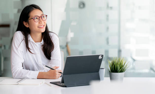 belle femme d’affaires réfléchie utilisant l’ordinateur, souriant et regardant au loin en réfléchissant à une entreprise prospère, en créant une nouvelle idée ou en se reposant distraite de la dure journée de travail au bureau. - thai ethnicity photos et images de collection