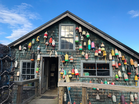 A shingled cottage in Maine with lobster buoys hanging off of it with lobster traps nearby; close up