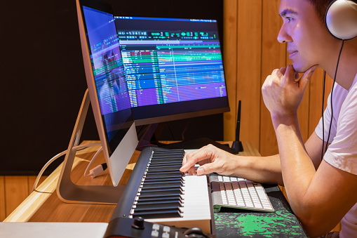 asian music producer arranging a song on midi keyboard and desktop computer in home studio. music production technology concept