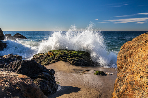 Wave breaking on rock in Malibu, California. Beach and rocks in foreground; Blue sky, Pacific ocean in the distance.
