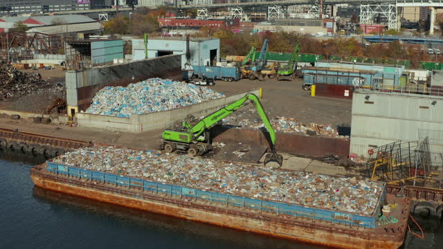 Aerial ascending footage of green loader standing on waterfront near barge loaded with plastic waste. Recycling facility. New York City, USA