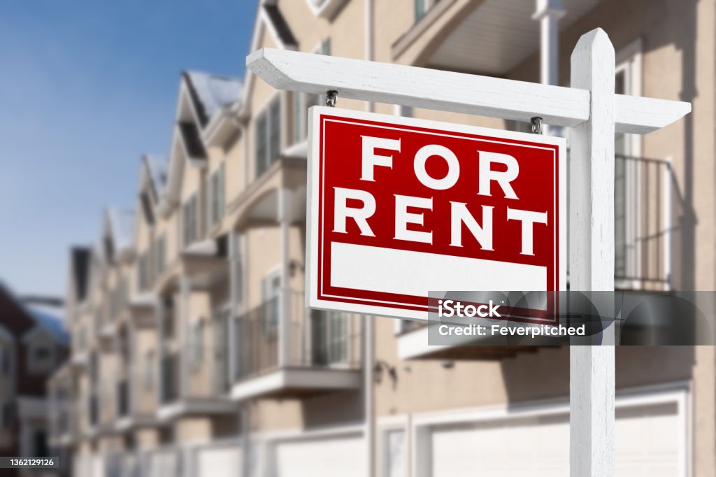 For Rent Real Estate Sign In Front of a Row of Apartment Condominiums Balconies and Garage Doors. Renting Stock Photo