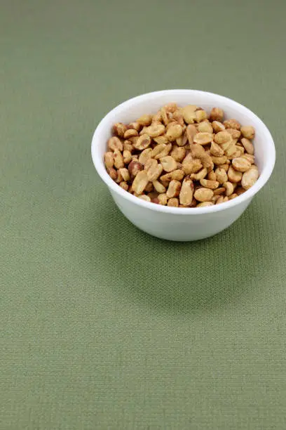 White round bowl of seasoned dry roasted peanuts with some peanut skins on a green table cloth. Snack food of dry roasted seasoned peanuts with sea salt and seasonings in a round white bowl close-up.