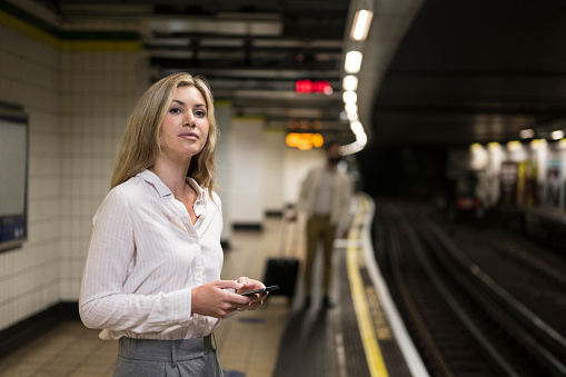 Waist-up view of Caucasian woman in summertime workwear standing on subway platform, holding smart phone, and looking down track with anticipation.