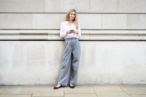 Full length front view of early 30s woman with long blond hair wearing summertime work attire and pausing from checking smart phone to smile at camera.
