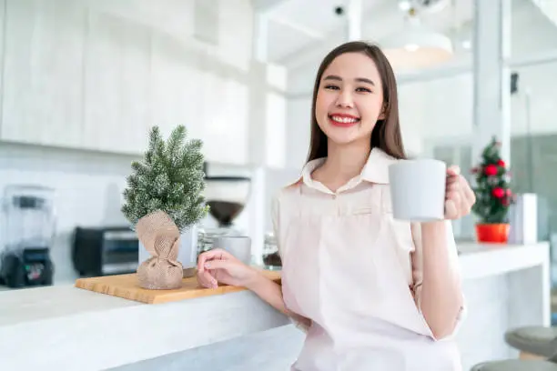 Happiness cheerful Asian female woman in uniform small business coffeeshop owner working in coffeeshop,chinese female barista say hi greeting hand gesture to camera welcome smiling and joyful Keep a Positive Attitude at Work Concepts.