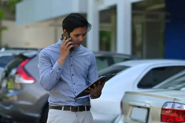 Photo of an insurance agent talking on mobile-phone while standing and holding a clipboard at the car park.