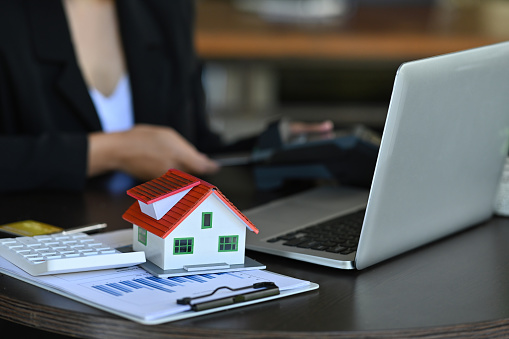 Photo of a working table surrounded by a computer laptop, paperwork, calculator, and house model.