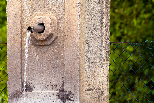 Old stone water fountain, spurt, drinking water free of charge in townsquare, green vegetation in the background. Galicia, Spain.