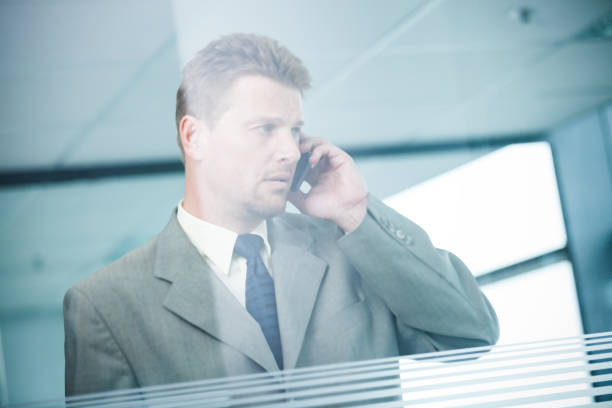 Serious businessman talking on the smart phone in office standing near big window. A view through the glass wall with white stripes.