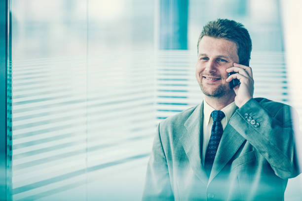 Happy businessman talking on the smart phone standing near big window. A view through the glass wall with white stripes.