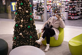 Pretty customer in mask, cozy white winter coat and high boots is sitting relaxed in armchair in shopping center near Christmas tree. Shopping mall . New normal concept. New Year's mood.