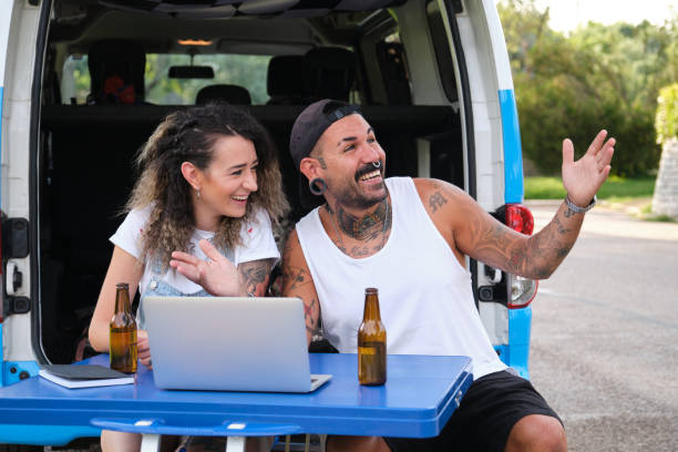 Young tattooed couple smile and talk in a laptop video call during a van trip. Young tattooed couple smiling and showing the place to heir family and friends in a video call using the laptop, sitting in a picnic table during a van trip. septum piercing stock pictures, royalty-free photos & images