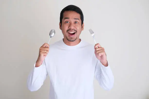Excited Asian man holding spoon and fork in his hands