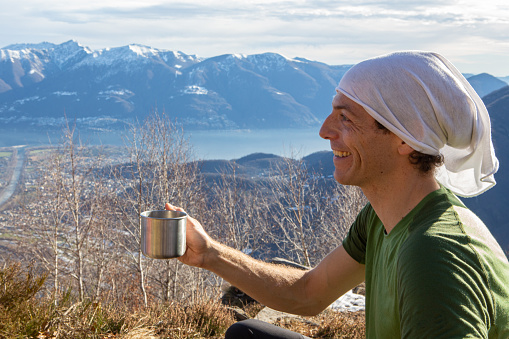 Young man hiker drinks from metal cup, mountain range