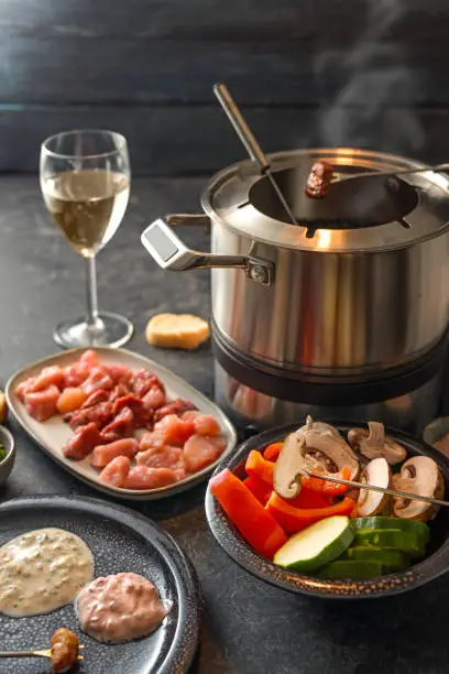 Fondue from vegetables and meat cooked at the table in a pot with boiling broth or hot oil, served on a festive dinner with dip sauce, bread and wine, dark gray background, selected focus, narrow depth of field