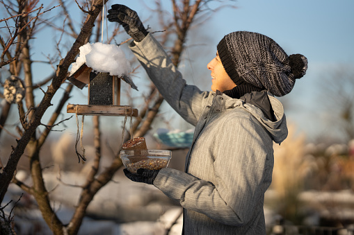 Happy woman in warm winter clothes refilling birds feeder in her garden, side view, upper body