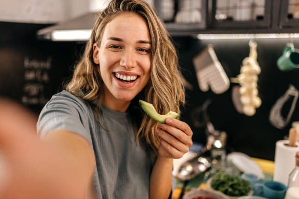 Woman eating avocado and taking selfie Portrait of smiling young women enjoying healthy slice of avocado during morning breakfast and taking selfie selfie stock pictures, royalty-free photos & images