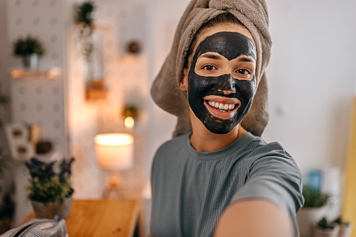 Portrait of a young smiling beautiful woman with towel on head and charcoal black mask taking a selfie