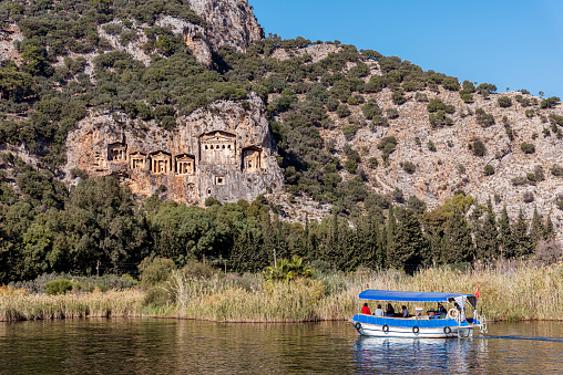 Koycegiz, Mugla, Turkey - November 21, 2021: In the background is the rock tombs of the kings in the ancient city of Kaunos and in the foreground the tourboat taking the tourists around. Rock tombs of the kings at ancient city of Kaunos, a Unesco World Heritage Site at Dalyan, Koycegiz, Mugla Province, Turkey. In the foreground, boats left next to the reeds by the river can be seen.