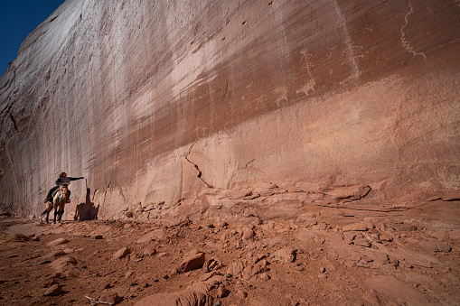 Navajo Boy riding horse along a sandstone red cliff with petroglyphs at the Monument Valley Tribal Park on a sunny day